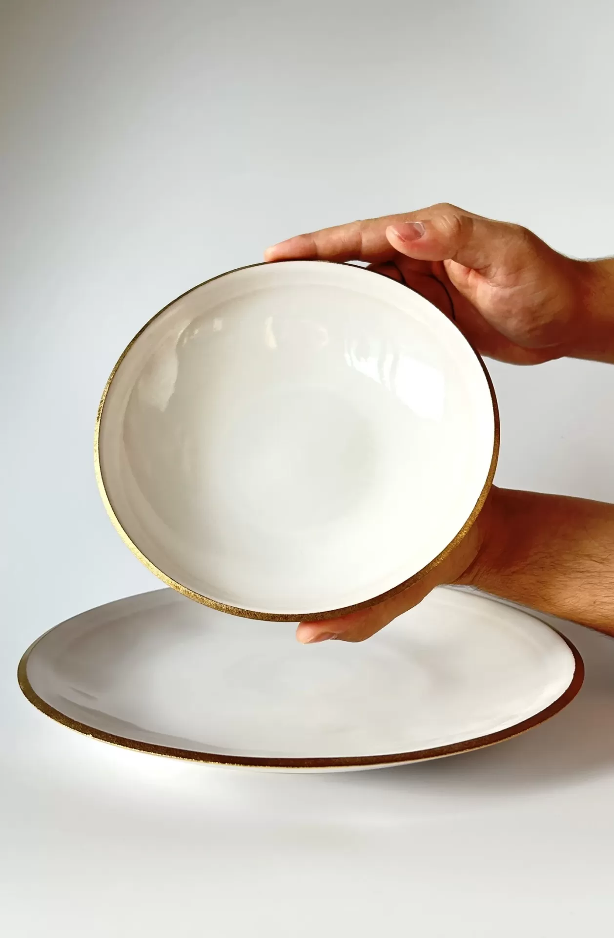 Conjunto de pratos branco com bordas douradas em fundo branco, uma peça segurada por mãos.