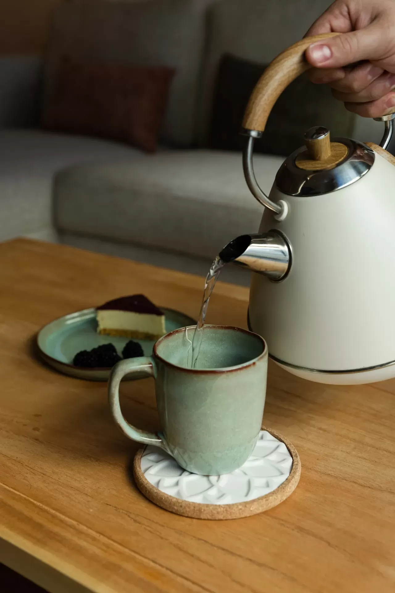 Bule metálico branco com detalhes em madeira a verter água numa caneca verde sobre mesa de madeira com bolo e amoras num prato verde