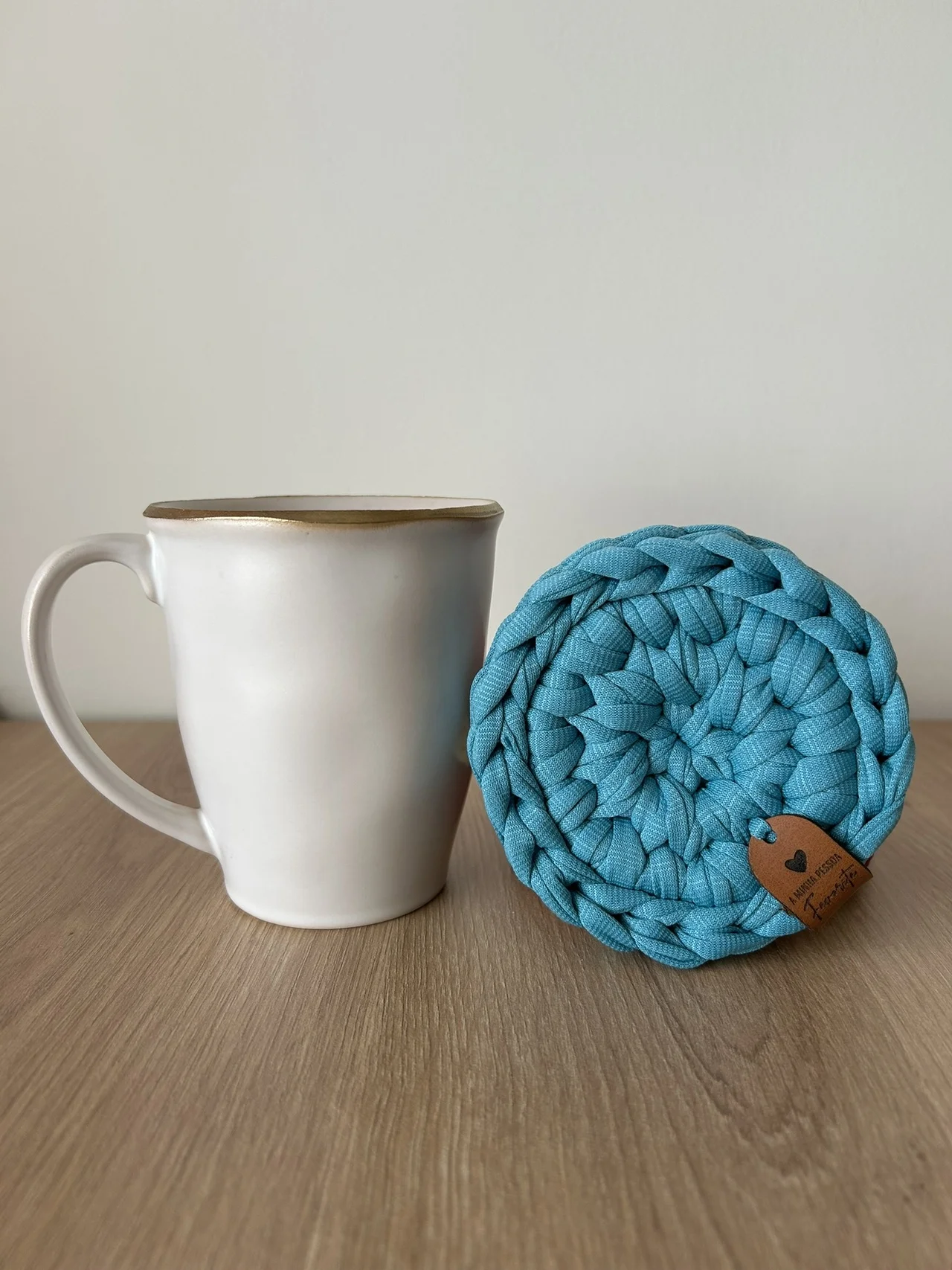 Caneca branca com borda castanha e descanso de copos azul trançado em tecido sobre mesa de madeira.