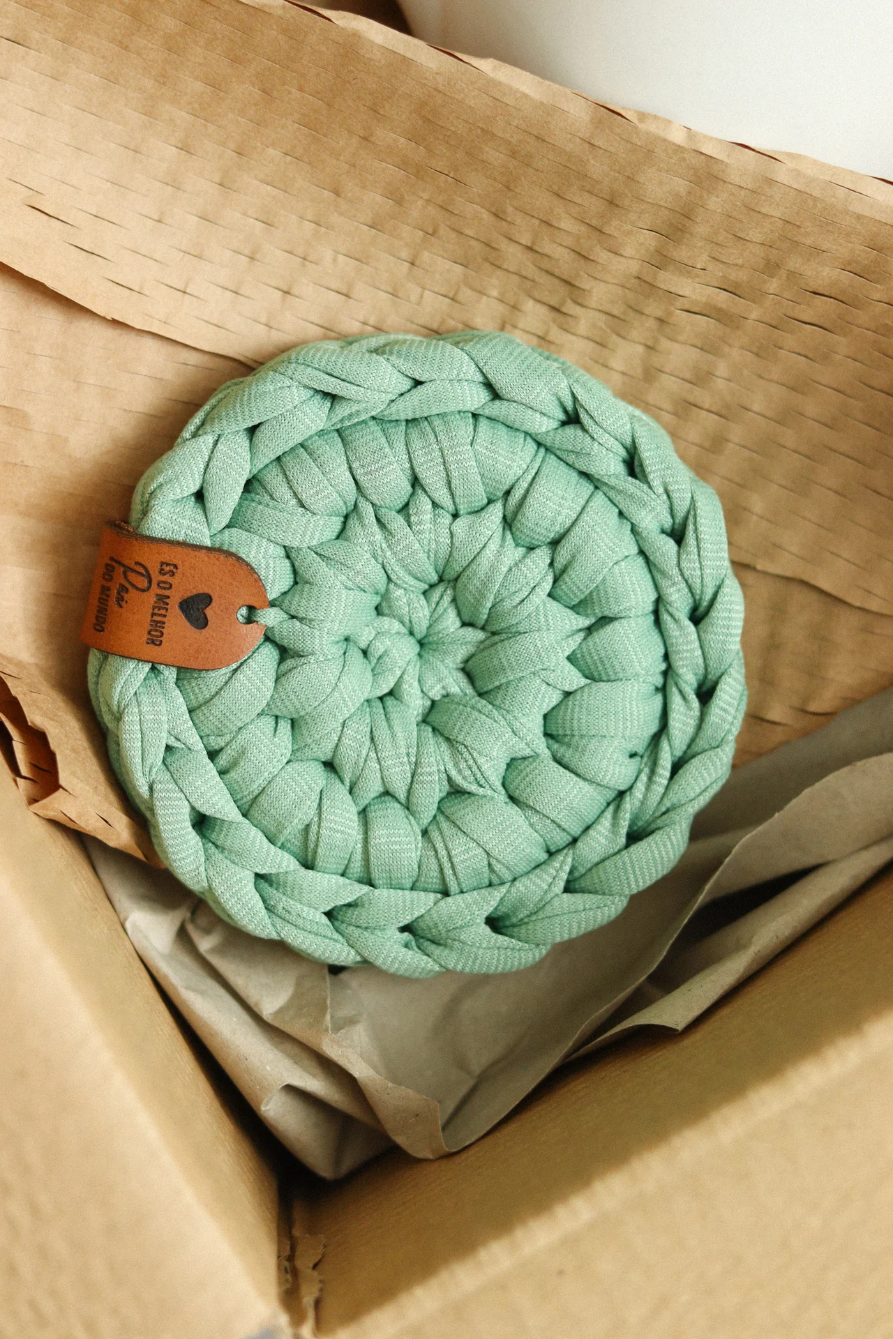 Prato raso redondo verde água tecido, com etiqueta em pele e embalagem de papel kraft.