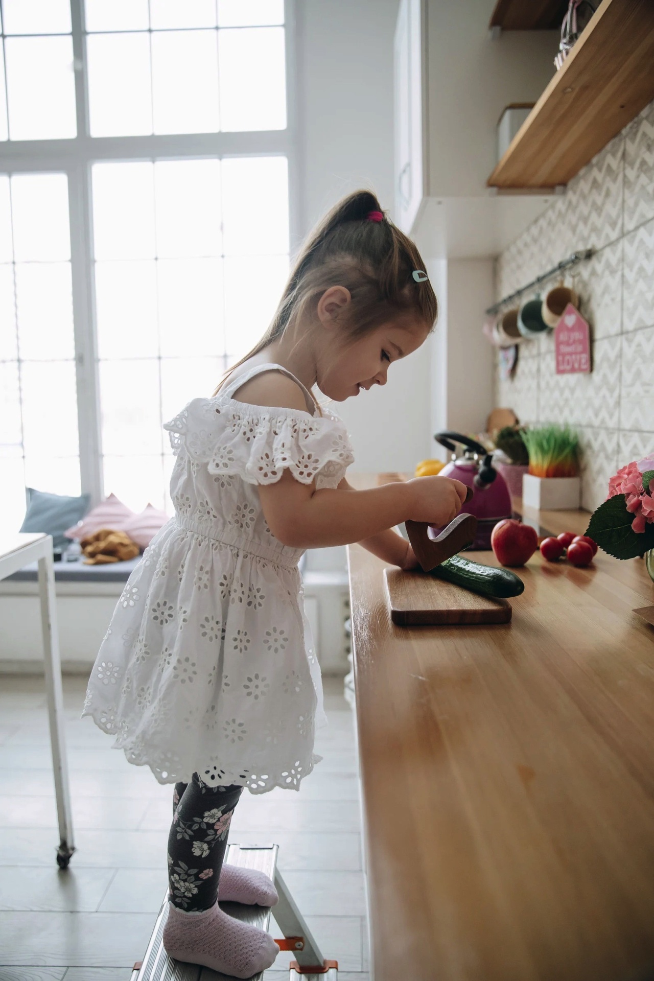 Menina com vestido branco rendado e leggings pretas a cozinhar na cozinha com bancada de madeira e parede de azulejos cinzentos