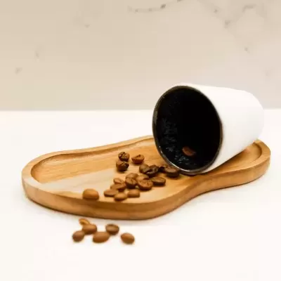 Copo de cerâmica branco com interior preto em bandeja de madeira com grãos de café.