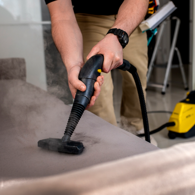 Máquina a vapor amarela e preta a limpar sofá cinzento com mangueira preta.