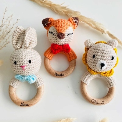 Tres mordedores de madera con figuras de crochet de conejo, zorro y león sobre superficie blanca