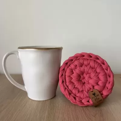 Caneca branca com borda dourada ao lado de suporte trançado rosa com etiqueta em pele numa mesa de madeira