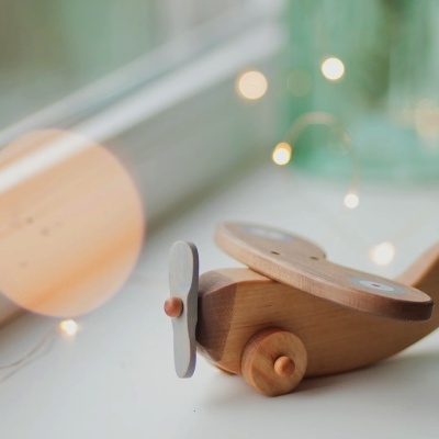 Avião de madeira clara com hélice e rodas numa superfície branca junto à janela.