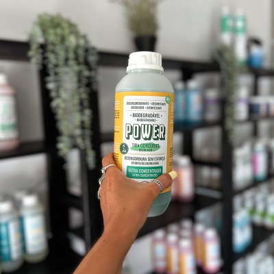 Frasco de produto desengordurante biodegradável com rótulo verde e amarelo sendo segurado por mão, fundo com prateleiras cheias de frascos.