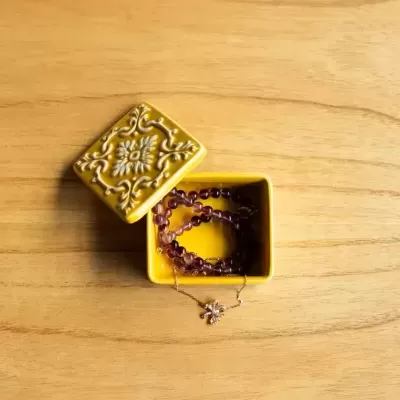 Caixa quadrada amarela decorativa com colares dentro, sobre superfície de madeira clara.