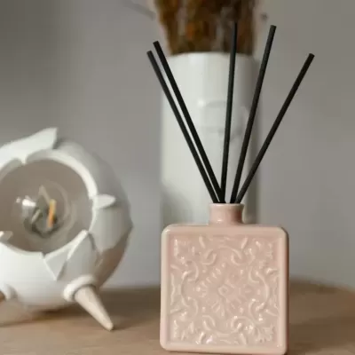 Difusor de ambientador rosa com varetas pretas sobre madeira com fundo de vaso branco e ornamento