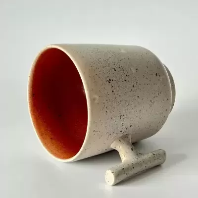 Caneca de cerâmica bege com interior vermelho alaranjado deitada sobre fundo branco