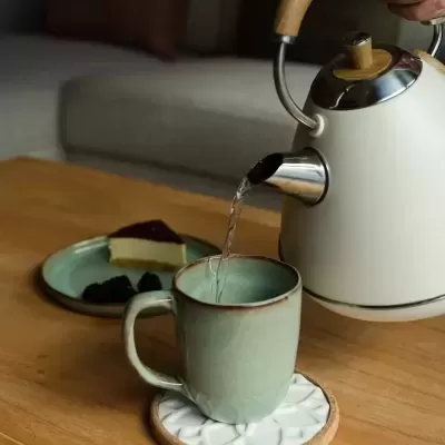 Bule metálico branco com detalhes em madeira a verter água numa caneca verde sobre mesa de madeira com bolo e amoras num prato verde