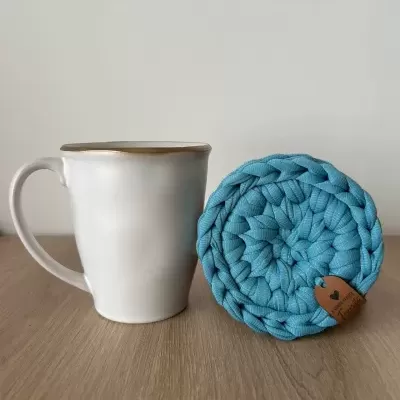 Caneca branca com borda castanha e descanso de copos azul trançado em tecido sobre mesa de madeira.