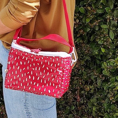 Bolsa vermelha pequena com padrão branco, fecho e alça, com fundo de folhagem verde