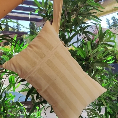 Almofada pequena bege com listras pendurada em mão, plantas ao fundo