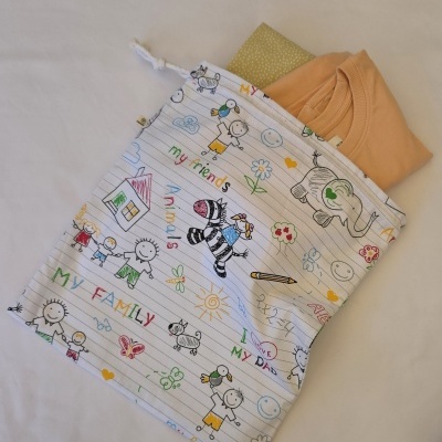 Saco de pano branco com desenhos infantis coloridos e uma t-shirt bege dobrada por cima