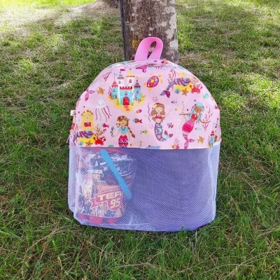 Mochila infantil cor-de-rosa com padrão colorido de sereias e castelos junto a uma árvore sobre relvado