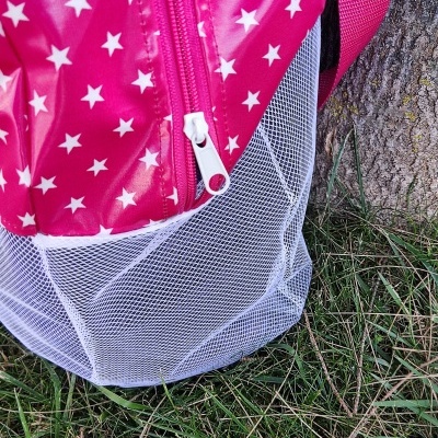 Mochila rosa com estrelas brancas e parte inferior em tela branca junto a uma árvore na relva