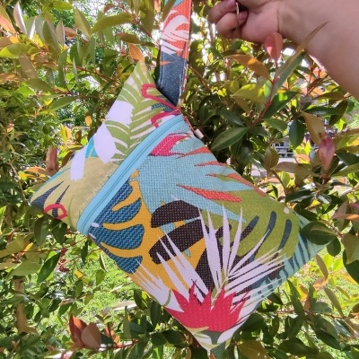 Bolsa pequena colorida com padrão tropical, zíper azul claro e alça para pulso segurada por mão ao ar livre