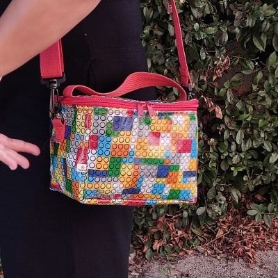 Mala tiracolo colorida com padrão de círculos, usada por mulher com vestido preto, ao ar livre.