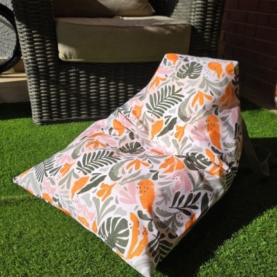 Pufe de jardim estampado com folhas verdes, laranja e rosa, em relva artificial junto a cadeira de vime castanho com almofadas bege