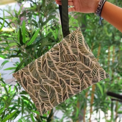 Bolsa com padrão de folhas verde e branco com alça preta, em frente a plantas verdes.