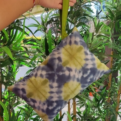 Mala de mão azul e amarela em tecido tie-dye com alça verde, segurada à frente de plantas verdes