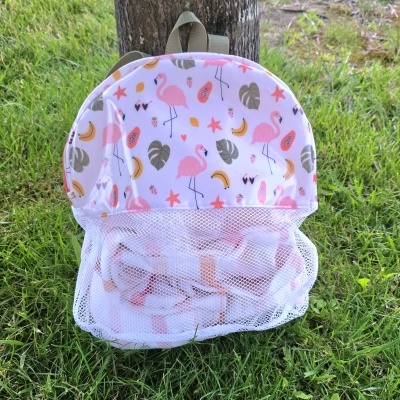 Mochila infantil branca com padrão de flamingos, folhas e frutas coloridas, pendurada em árvore na relva