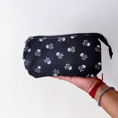 Necessaire preta com padrão de caveiras brancas segurada por uma mão com pulseiras