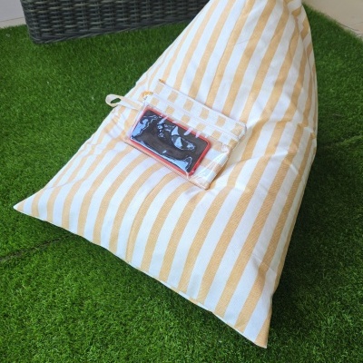 Pufe amarelo e branco riscado com bolso transparente contendo telemóvel, sobre relva artificial junto a cadeira de vime cinza com almofada bege.