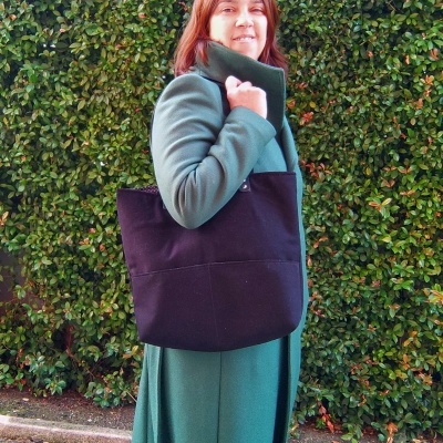 Mulher com casaco verde e mala preta sobre o ombro em frente a sebes verdes.