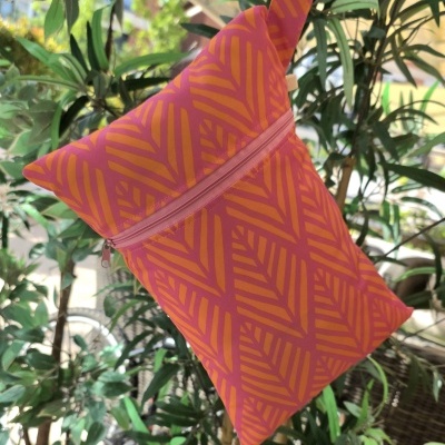 Bolsa de tecido rosa com padrão laranja e fecho de correr, pendurada entre plantas.