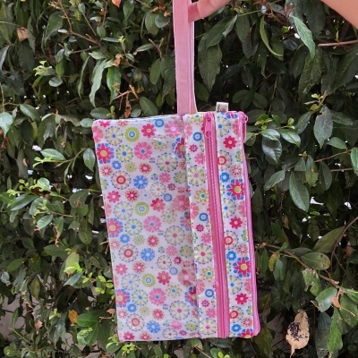 Bolsa floral colorida com fecho e alça rosa segurada por uma mão em fundo de folhagem