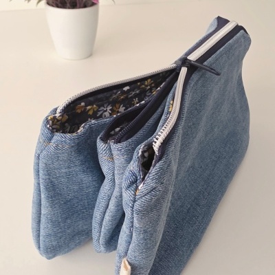 Bolsa azul imitando ganga com fecho zip em superfície branca junto a vaso com flores cor de rosa