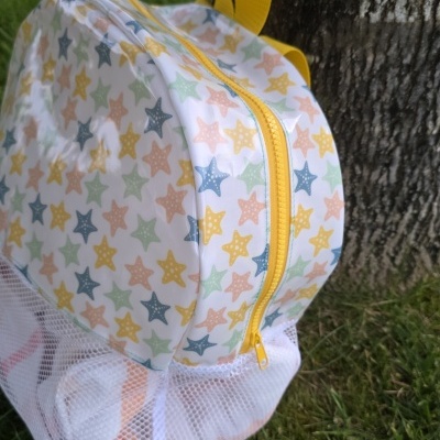 Mochila infantil com padrão colorido de estrelas e fecho amarelo pendurada num tronco de árvore.