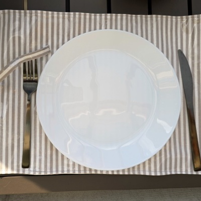 Conjunto de jantar com prato branco, talheres de metal e individual às riscas bege e branco sobre mesa de madeira.