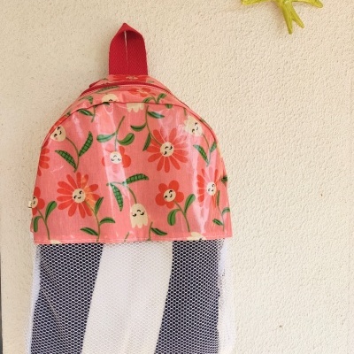 Mochila cor-de-rosa com flores e parte inferior em rede transparente e preta pendurada numa parede bege com um pássaro amarelo