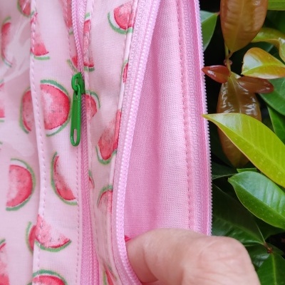 Bolsa cor-de-rosa com padrão de fatias de melancia e fecho verde, segura por mão com unhas vermelhas, folhas verdes ao fundo