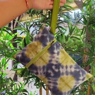 Bolsa tie-dye com fecho e alça verdes segurada por uma mão junto a plantas