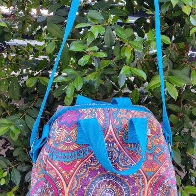 Mala de ombro colorida com padrão floral e mandala, alças azuis, pendurada numa planta