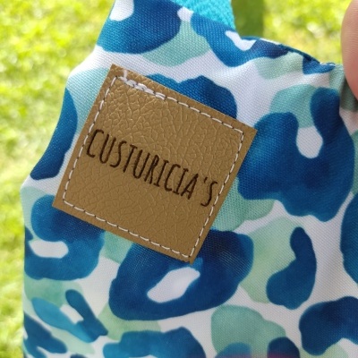 Produto tecido azul e verde com etiqueta de couro sintético castanho com texto 'CUSTURICIA'S'