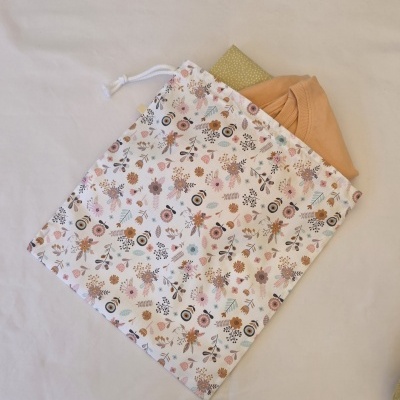 Saco de algodão florido com cordão branco e roupa laranja e verde dentro sobre fundo branco.