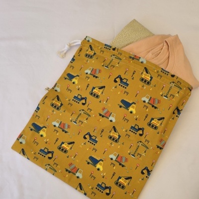 Saco de tecido amarelo com padrão de veículos de construção e cordão branco, com roupa e pano dentro