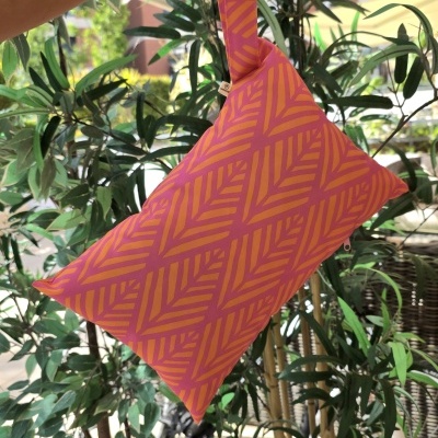 Bolsa de tecido rosa com padrão geométrico laranja e alça, segurada por uma mão, ao lado de plantas em vasos brancos.