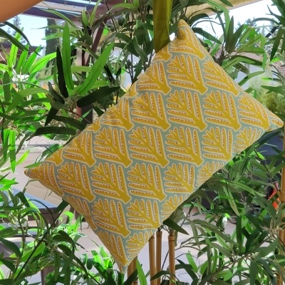Almofada retangular amarela com padrão e alça verde suspensa por mão em ambiente com plantas