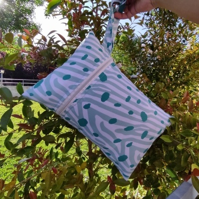 Bolsa pequena com padrão de riscas e manchas verdes, com fecho de correr e alça, segurada ao ar livre