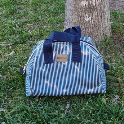 Mala de mão azul com riscas brancas e alças azuis no chão junto a árvore