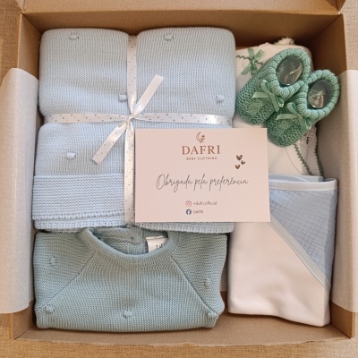 Conjunto para bebé com manta azul, camisola verde água, sapatinhos verdes e babygrow branco num caixa de cartão.