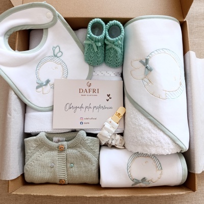 Conjunto de roupa e acessórios de bebé em caixa de cartão com detalhes em verde e branco, incluindo babete, botas, casaco, toalha e cartão de agradecimento