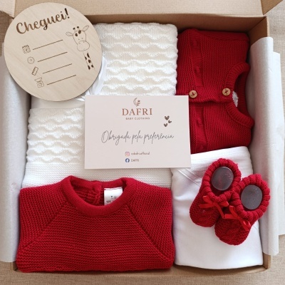 Caixa com roupa e acessórios de bebé em vermelho e branco, placa de madeira e cartão de agradecimento.