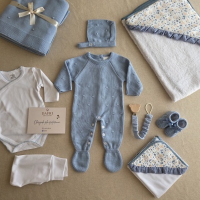 Conjunto completo para bebé em tons azul e branco com roupa, mantas e acessórios sobre fundo bege.